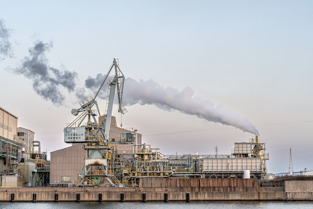 昼間の工場の様子と工場から出る煙が風に靡いて工場の上を流れています。